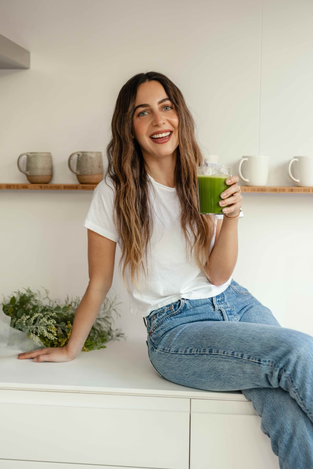 Meghan Swidler sitting on a kitchen counter, holding a glass of green juice and smiling