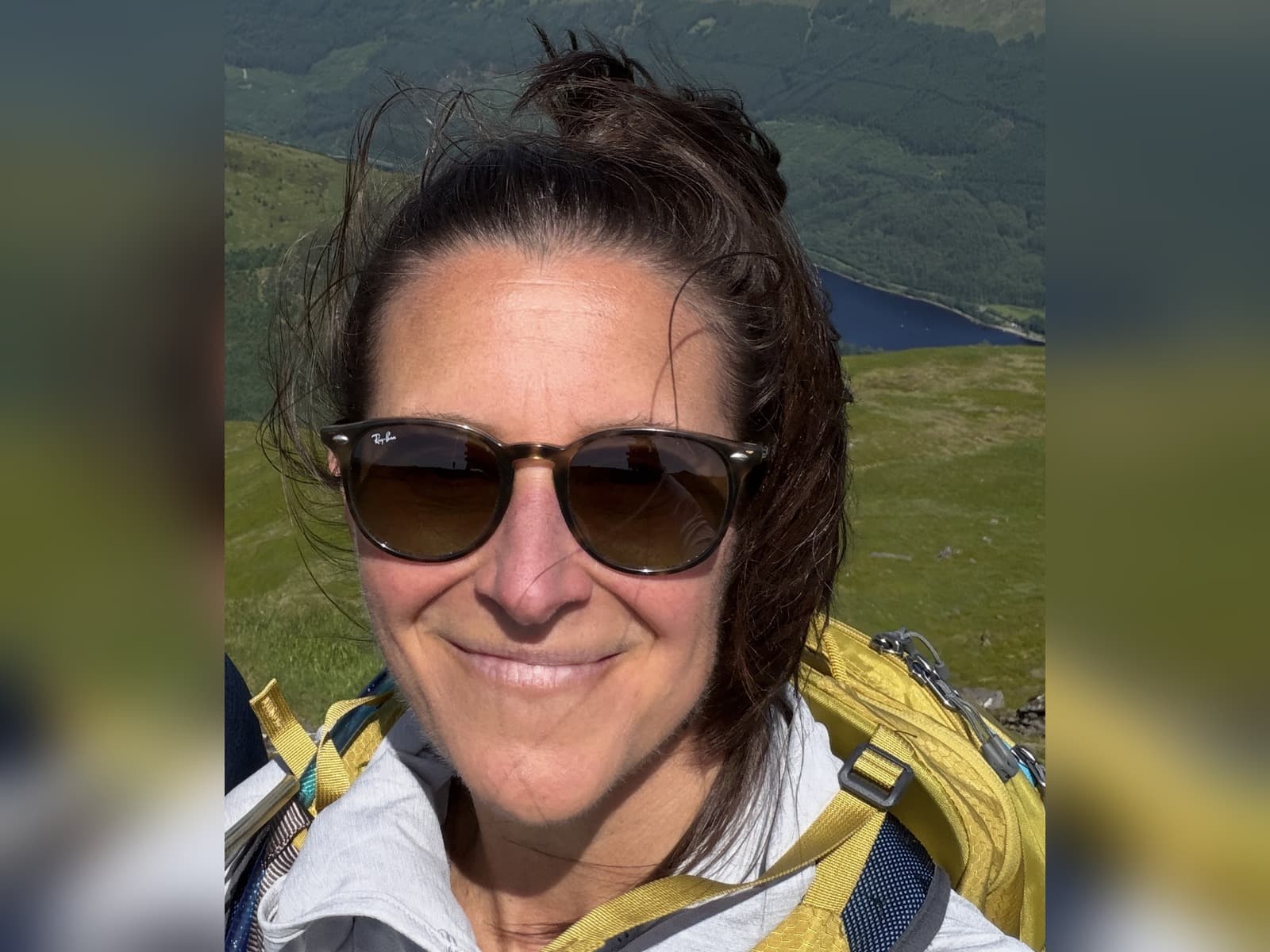 Suzanne, smiling on a hillside in sunglasses with a lake behind her